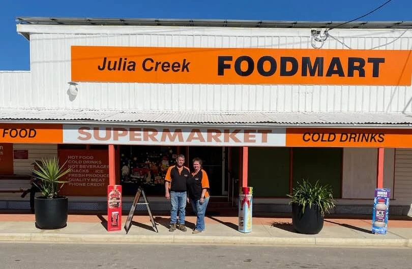 Julia Creek Foodmart shopfront: white corrugated iron building with the orange Foodmart sign, and two staff out front